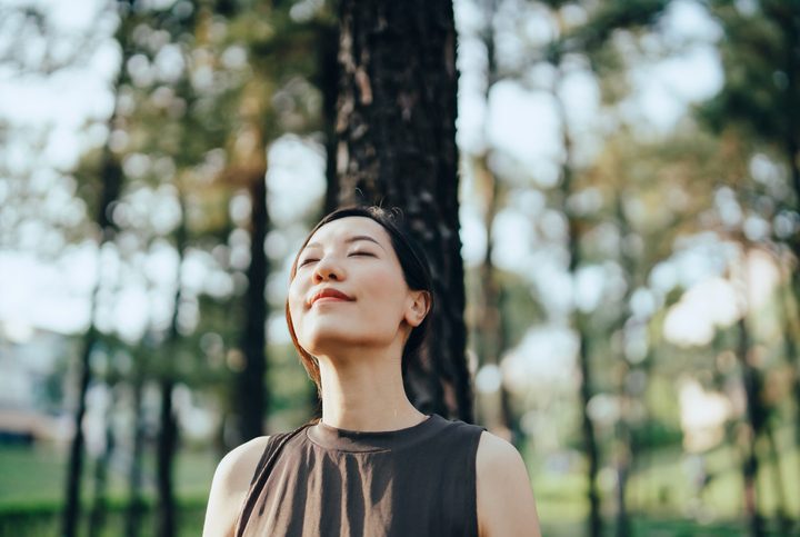 Young Asian woman with eyes closed leaning against tree in forest, with trees and sunbeam on background. Enjoying fresh air and connection with nature. Freedom in nature